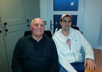 Two people sit indoors, smiling; left wearing a dark shirt, right in a white jacket and cap.