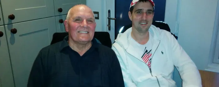 Two people sit indoors, smiling; left wearing a dark shirt, right in a white jacket and cap.