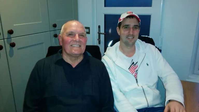 Two people sit indoors, smiling; left wearing a dark shirt, right in a white jacket and cap.