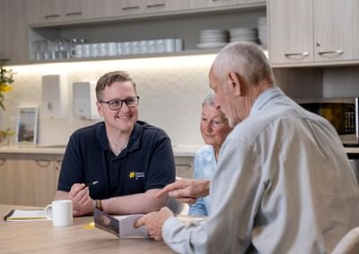 Three people sit at a kitchen table, a staff member in a polo with Cancer Council logo, smiling.