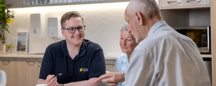 Three people sit at a kitchen table, a staff member in a polo with Cancer Council logo, smiling.