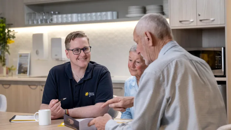 Three people sit at a kitchen table, a staff member in a polo with Cancer Council logo, smiling.