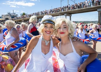 Two people in white dresses with blonde wigs smile on a sunny beach; crowd on a pier.