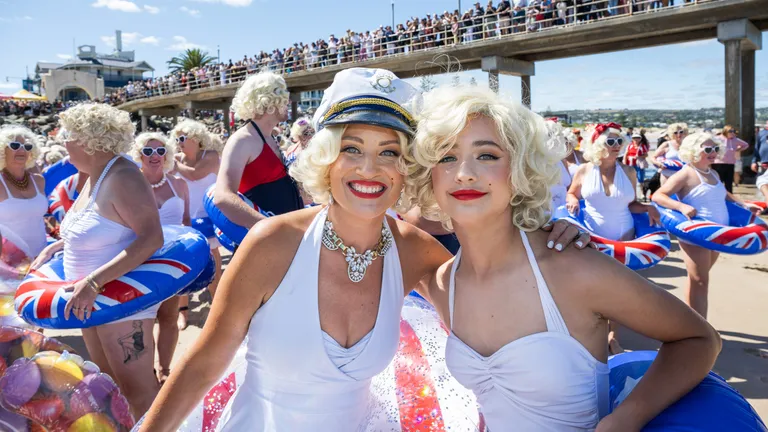 Two people in white dresses with blonde wigs smile on a sunny beach; crowd on a pier.