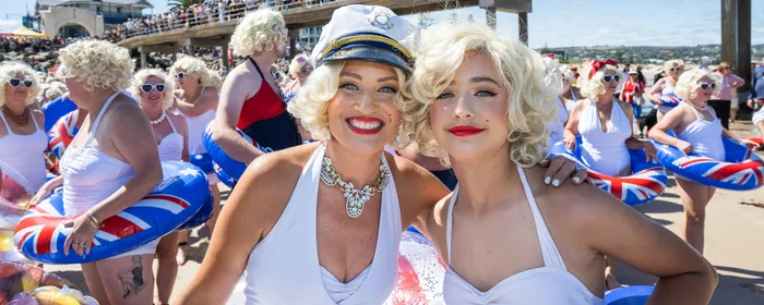 Two people in white dresses with blonde wigs smile on a sunny beach; crowd on a pier.
