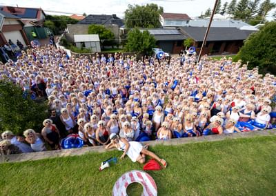 Large crowd in white outfits and sunglasses in a suburban street; a person lies on the grass in front.