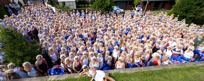 Large crowd in white outfits and sunglasses in a suburban street; a person lies on the grass in front.