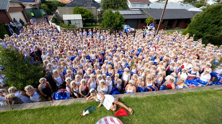 Large crowd in white outfits and sunglasses in a suburban street; a person lies on the grass in front.