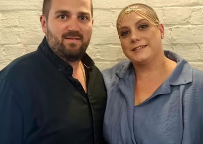 Two people pose together against a white brick wall; one in black, the other in blue with a gold headpiece.
