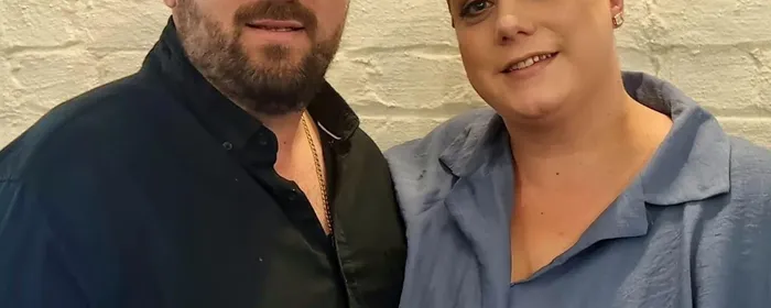 Two people pose together against a white brick wall; one in black, the other in blue with a gold headpiece.