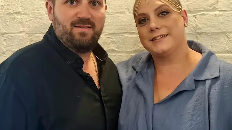 Two people pose together against a white brick wall; one in black, the other in blue with a gold headpiece.