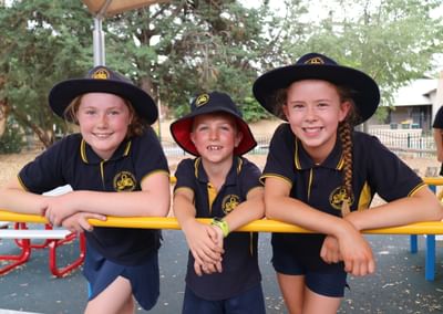 Three children in navy uniforms with hats lean on a yellow railing at a playground, smiling.