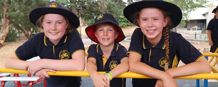 Three children in navy uniforms with hats lean on a yellow railing at a playground, smiling.