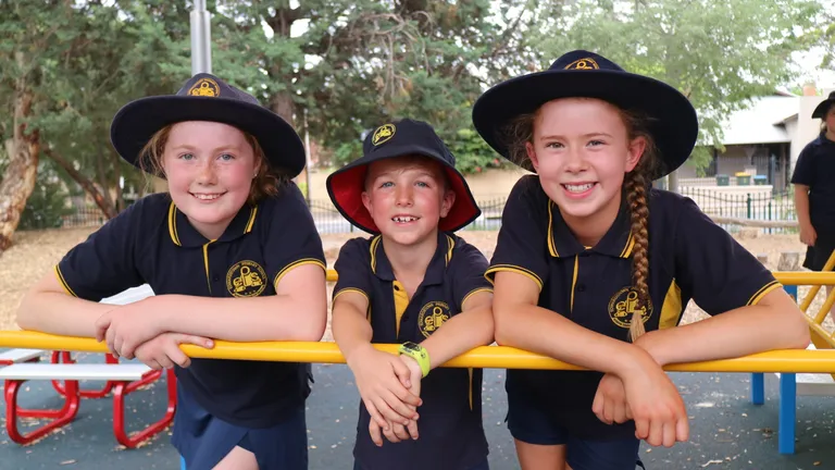 Three children in navy uniforms with hats lean on a yellow railing at a playground, smiling.