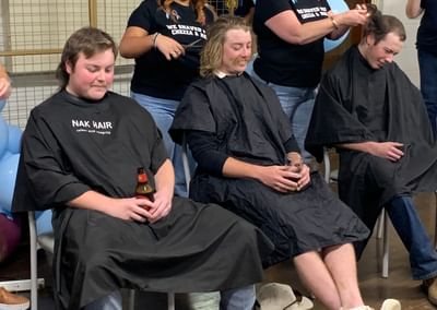 Three people in barber capes sit while two stylists cut hair at a balloon‑decorated indoor event.