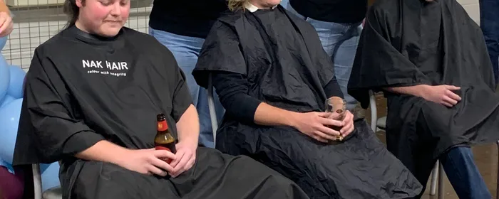 Three people in barber capes sit while two stylists cut hair at a balloon‑decorated indoor event.
