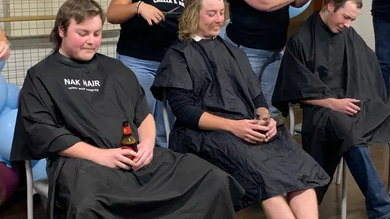 Three people in barber capes sit while two stylists cut hair at a balloon‑decorated indoor event.