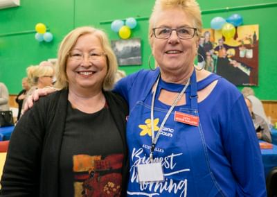 Two people smiling and posing together at a community event, indoors with green walls and balloons.