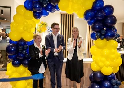 Three people applaud under a blue and yellow balloon arch at an indoor reception.
