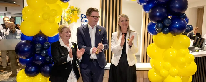 Three people applaud under a blue and yellow balloon arch at an indoor reception.