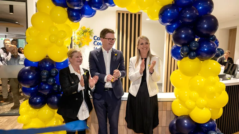 Three people applaud under a blue and yellow balloon arch at an indoor reception.