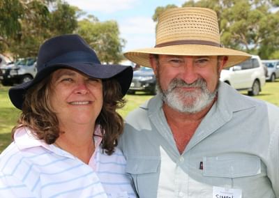 Two people outdoors wearing wide-brimmed hats, smiling, in a sunny park with cars in the background.