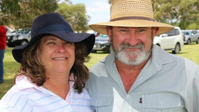 Two people outdoors wearing wide-brimmed hats, smiling, in a sunny park with cars in the background.