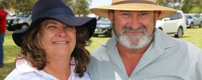 Two people outdoors wearing wide-brimmed hats, smiling, in a sunny park with cars in the background.
