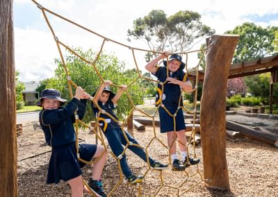 Three children in navy uniforms and sun hats climb a rope net between wooden posts at a sunny playground.