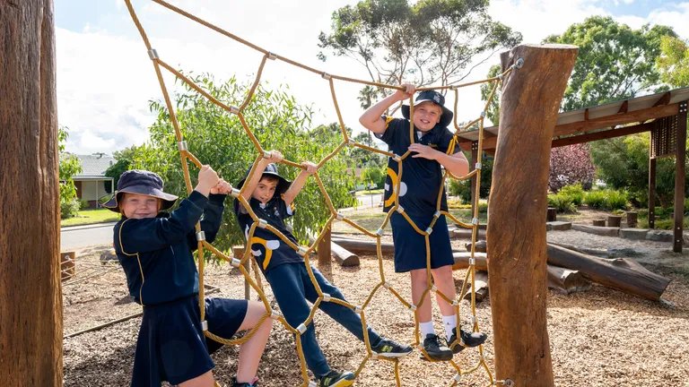 Three children in navy uniforms and sun hats climb a rope net between wooden posts at a sunny playground.