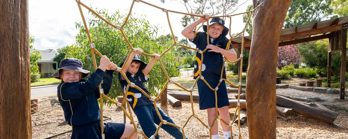 Three children in navy uniforms and sun hats climb a rope net between wooden posts at a sunny playground.