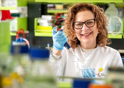 Lab worker with curly hair and glasses, in a white coat, using a pipette in a bright lab.