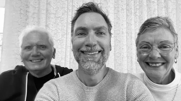 Three adults smiling at the camera indoors, with lace curtains behind them.
