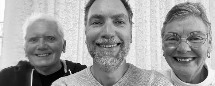 Three adults smiling at the camera indoors, with lace curtains behind them.