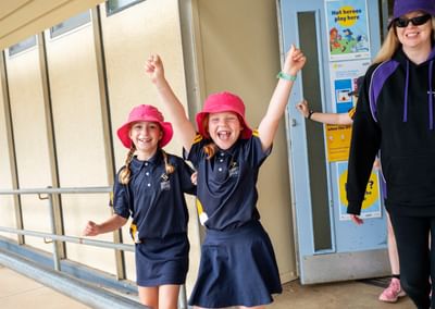 Two children in navy uniforms with pink hats raise their arms beside an adult in a purple hoodie.