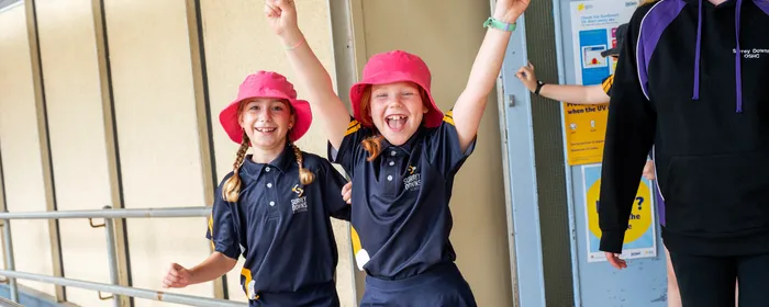 Two children in navy uniforms with pink hats raise their arms beside an adult in a purple hoodie.