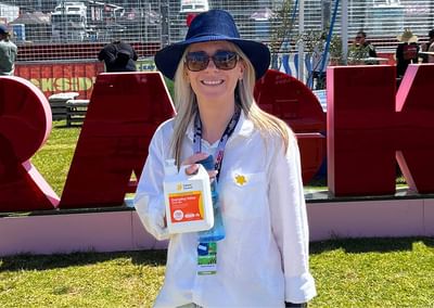 Person wearing white shirt and blue hat, smiling, holding sanitiser bottle at an outdoor event.