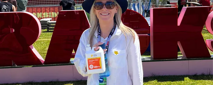 Person wearing white shirt and blue hat, smiling, holding sanitiser bottle at an outdoor event.