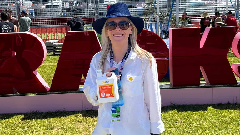 Person wearing white shirt and blue hat, smiling, holding sanitiser bottle at an outdoor event.