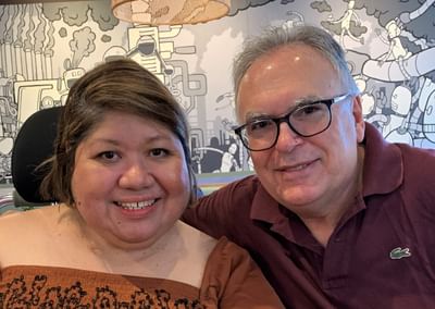 Two people smile at the camera in front of a mural; one wears an orange top, other a maroon polo.