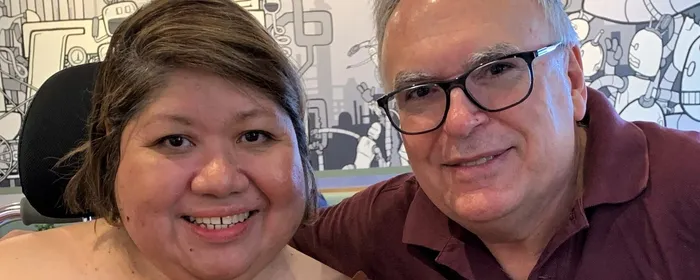 Two people smile at the camera in front of a mural; one wears an orange top, other a maroon polo.