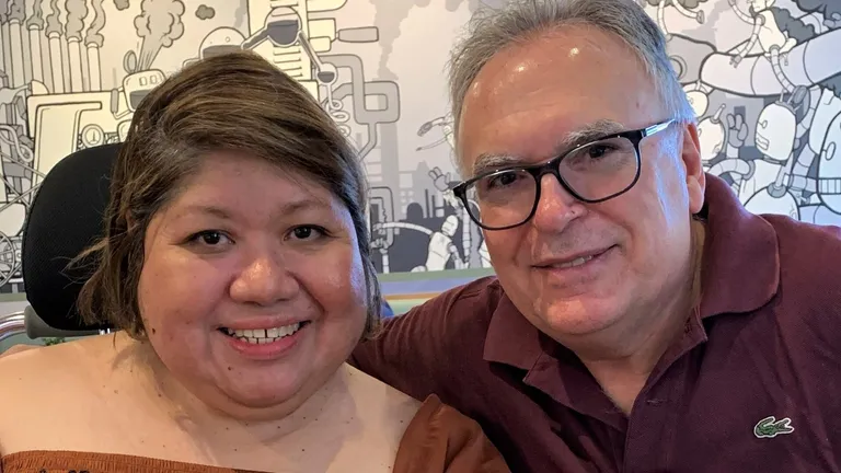 Two people smile at the camera in front of a mural; one wears an orange top, other a maroon polo.