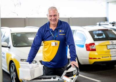 Smiling person in blue and yellow polo, pushing a brochure cart beside a Cancer Council vehicle.