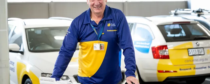 Smiling person in blue and yellow polo, pushing a brochure cart beside a Cancer Council vehicle.