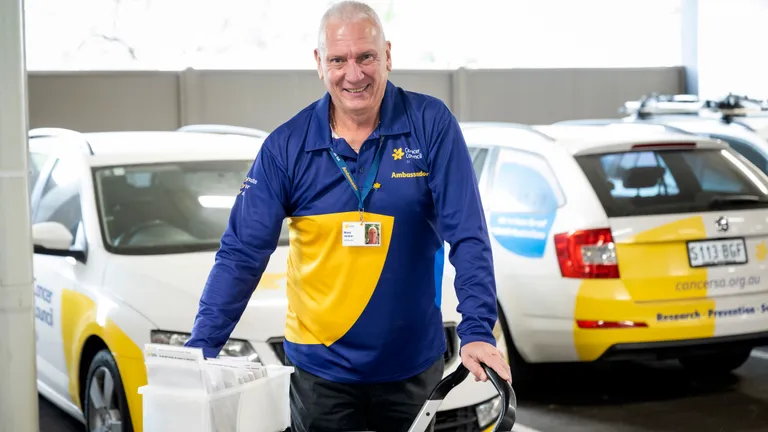 Smiling person in blue and yellow polo, pushing a brochure cart beside a Cancer Council vehicle.