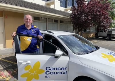 Person in blue and yellow polo by white Cancer Council SA car, purple-leaved tree in background.