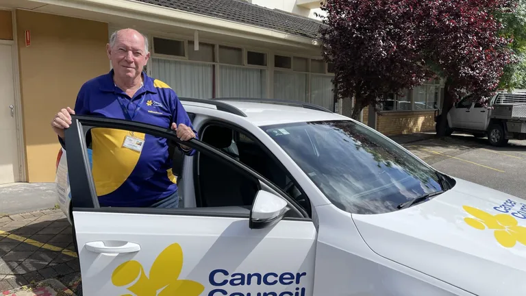 Person in blue and yellow polo by white Cancer Council SA car, purple-leaved tree in background.
