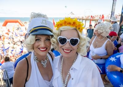 Two people with blonde wigs in white outfits at a crowded beach party; one wears a captain's hat.