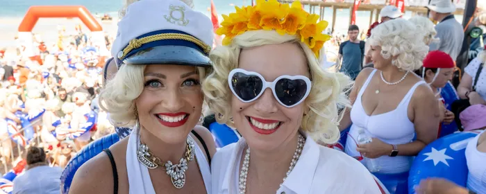 Two people with blonde wigs in white outfits at a crowded beach party; one wears a captain's hat.