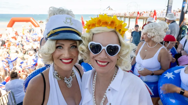 Two people with blonde wigs in white outfits at a crowded beach party; one wears a captain's hat.